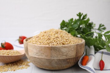 Raw bulgur in bowl, vegetables and parsley on table, closeup