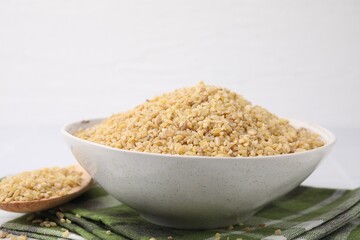 Bowl and spoon with raw bulgur on table against white background, closeup