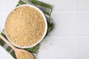 Bowl and spoon with raw bulgur on white tiled table, top view. Space for text