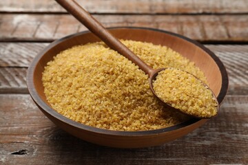 Bowl and spoon with raw bulgur on wooden table, closeup