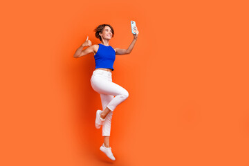Fototapeta na wymiar Full size profile photo of attractive sporty girl jump take selfie smart phone show thumb up empty space isolated on orange color background