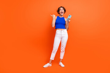 Full size photo of pretty impressed girl hold smart phone look direct finger empty space isolated on orange color background
