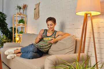 delighted veggie woman sitting on couch near lamp and eating fresh fruit salad on cozy sofa