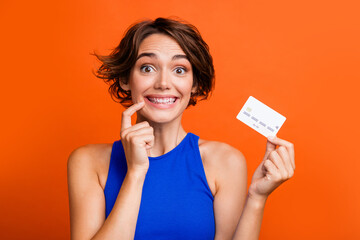 Photo of pretty cheerful girl finger touch lips toothy smile hand hold debit plastic card isolated on orange color background