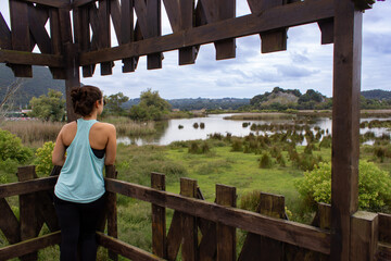 Young woman looking at a beautiful marsh after a run or a walk. long brown hair in a bun. She is wearing sports clothing and a sleeveless t-shirt and standing on a wooden platform. cloudy summer day.