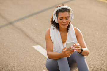 Athletic lady wearing headphones using fitness app on smartphone outside