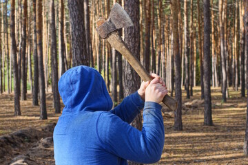 one male criminal in blue clothes and a hood swings hands a big axe aggressively on the street among the trees