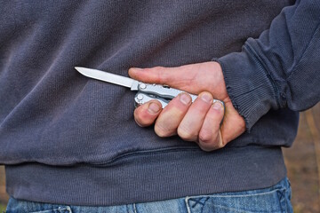 hand of a male criminal in black clothes and a gray knife behind his back on the street