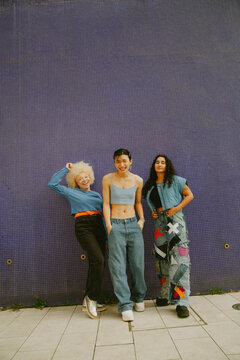 Three multiracial friends posing in cool outfits in front of blue wall smiling and looking into camera