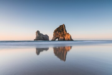 Serene beach scene featuring two large cliffs in the distance. Wharariki Beach, New Zealand - obrazy, fototapety, plakaty