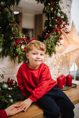 Happy child in red sweater hold hand mother. Smiling little boy near Christmas tree at home. New Year 2024, Merry Christmas. Сoncept of family holiday. Tree with garlands decorated interior of house.