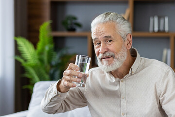Close-up portrait of an elderly gray-haired man sitting at home on the couch holding a glass of...
