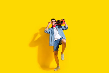 Full size photo of cool guy wear jeans jacket shorts touch sunglass hold boombox at summer party isolated on yellow color background