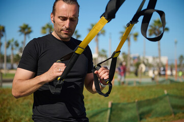 Determined muscular build young adult man, sportsman, athlete exercising with suspension straps...