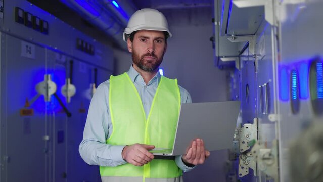 Professional electrical engineer in protective clothing using laptop at electrical substation. Man checking data and meter readings in technical room of factory looking at camera showing ok thumbs up.