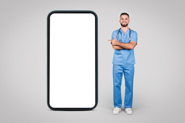 Male doctor in blue, pointing at huge phone screen, grey background