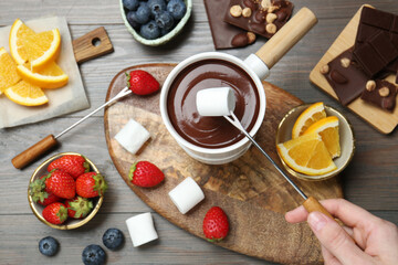 Woman dipping sweet marshmallow in fondue pot with melted chocolate at wooden table, top view