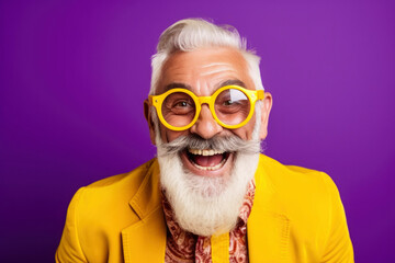 Portrait of cheerful elderly gray-haired bearded grandfather in funny sunglasses and bright extravagant clothes on plain purple background. Retired hipster, seniors party, carnival