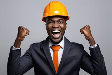Dark-skinned African American young male engineer in business suit and hardhat rejoices, smiles, raised his hands up. Concept of professional worker, architect, builder, manager, businessman