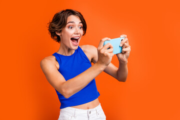 Photo of positive funky nice girl open mouth hands hold smart phone play games isolated on orange color background