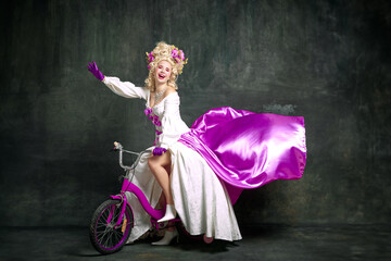 Cheerful charming woman in old-fashioned, aristocratic dress sitting, riding children's bike against vintage background.