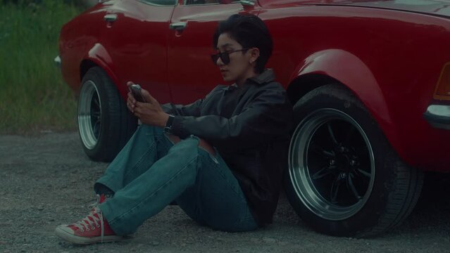 A brutal girl in a brown leather jacket sits outside her classic red car and texts on her phone