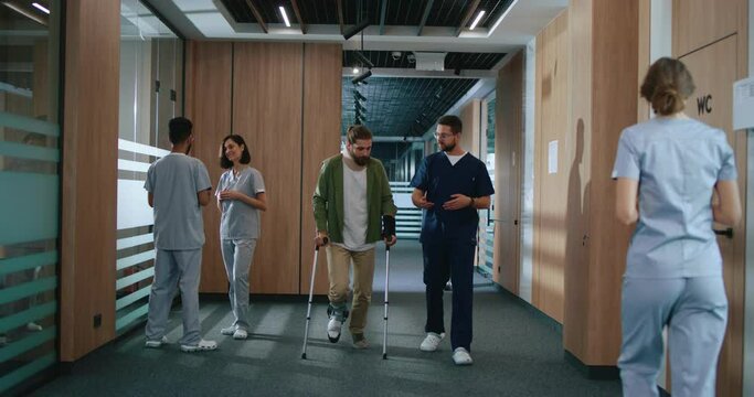 Caucasian kind physiotherapist supporting and talking with man using crutches. Medical workers spending time communicating and assisting people in contemporary hospital.