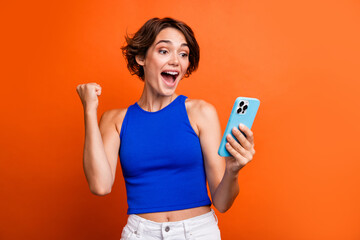 Photo of cheerful lucky girl dressed blue top rising fist winning game modern gadget isolated orange color background - obrazy, fototapety, plakaty
