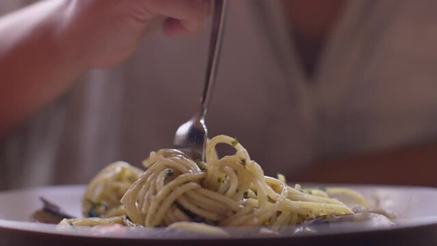 A young woman wraps spaghetti with vongole on a fork and eats it, curly long spaghetti from the plate goes into her mouth, a plate of spaghetti is eaten by a girl, pasta disappears from the plate