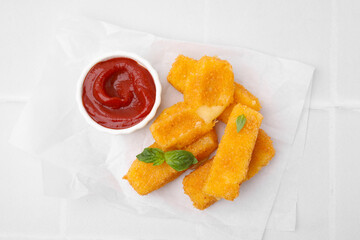 Tasty fried mozzarella sticks with basil leaves and ketchup on white tiled table, top view