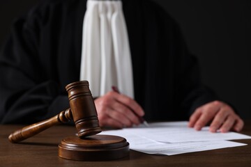 Judge with gavel writing in papers at wooden table against black background, closeup