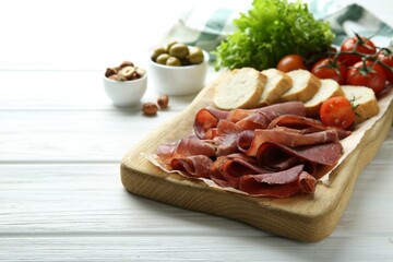 Board with delicious bresaola served with bread and tomato on white wooden table. Space for text