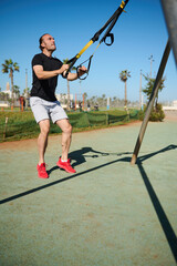 Young adult male athlete exercising with suspension straps, doing jumping exercises outdoors
