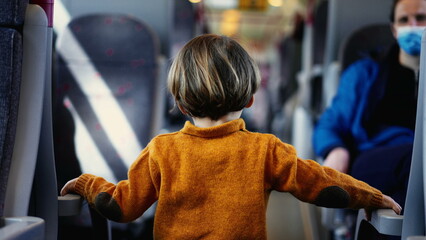 Young boy in a sunny yellow pullover navigates the train corridor, hands grasping the seat...