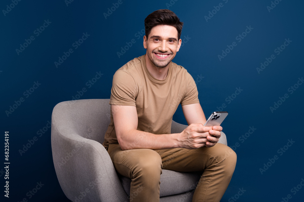 Poster Photo of toothy beaming intelligent man dressed beige t-shirt sit in armchair with smartpone in arms isolated on blue color background