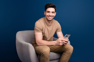 Photo of toothy beaming intelligent man dressed beige t-shirt sit in armchair with smartpone in arms isolated on blue color background