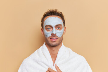 Close-up of young blue-eyed man with grey clay facial mask inside peach color studio, spa...