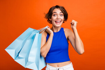 Photo of lovely delighted lady hold store bags raise fist success achievement isolated on orange...