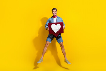 Fototapeta na wymiar Full size photo of overjoyed guy wear jeans jacket shorts jumping hold heart tap like on comment isolated on yellow color background