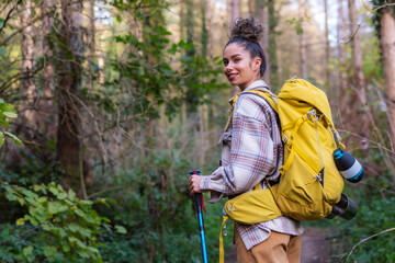 A young woman with curly hair in her 20s exploring the forest trail during autumn, wearing warm clothing and equipped with a yellow backpack, trekking poles, and binoculars.