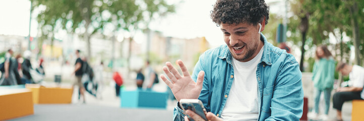 Clouseup, young bearded man in denim shirt sitting in wireless headphones making video call on mobile phone on busy street modern city background. - obrazy, fototapety, plakaty