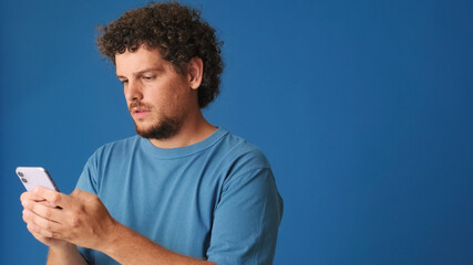 Pensive guy with curly hair dressed in blue t-shirt, holds mobile phone in his hands c space for...