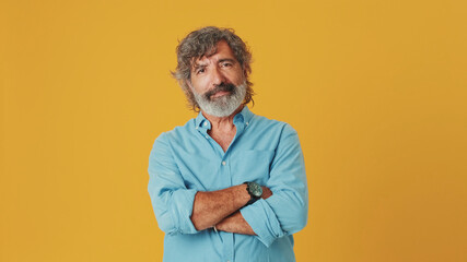 Elderly white-haired bearded man wears a blue shirt, looks at the camera with his arms crossed,...