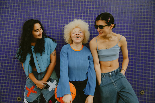 Three multiracial sporty friends in cool outfits posing with basketball in front of blue wall outdoors laughing