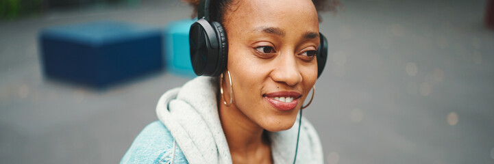 Close-up cute African girl with ponytail wearing denim jacket, in crop top with national pattern, listening to music with headphones in modern building background.