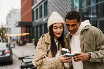 Smiling couple using smartphone and drinking coffee while standing at street