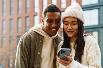 Beautiful couple smiling and looking at smartphone screen while standing at street