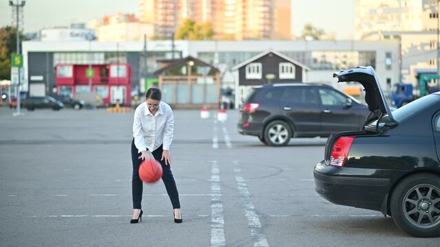a young woman in formal office clothes plays with a basketball in the parking lot and throws the ball into the trunk of a sedan