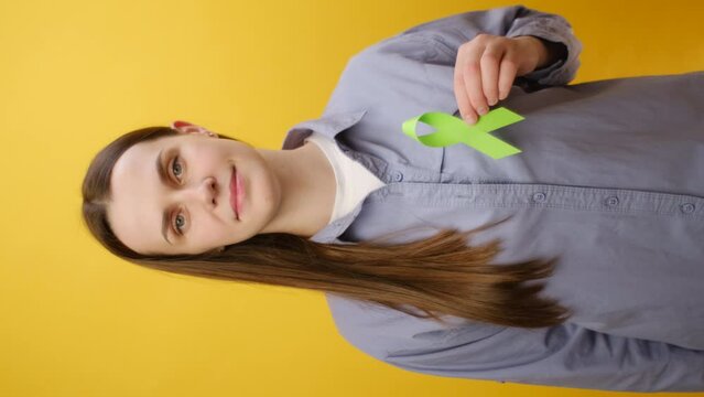 Vertical video of young woman holding small lime green ribbon, posing isolated over yellow studio background. Mental health awareness, world mental health day, world kidney day, organ donation concept