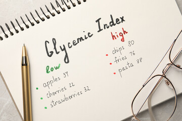 List with products of low and high glycemic index in notebook, pen and glasses on table, top view
